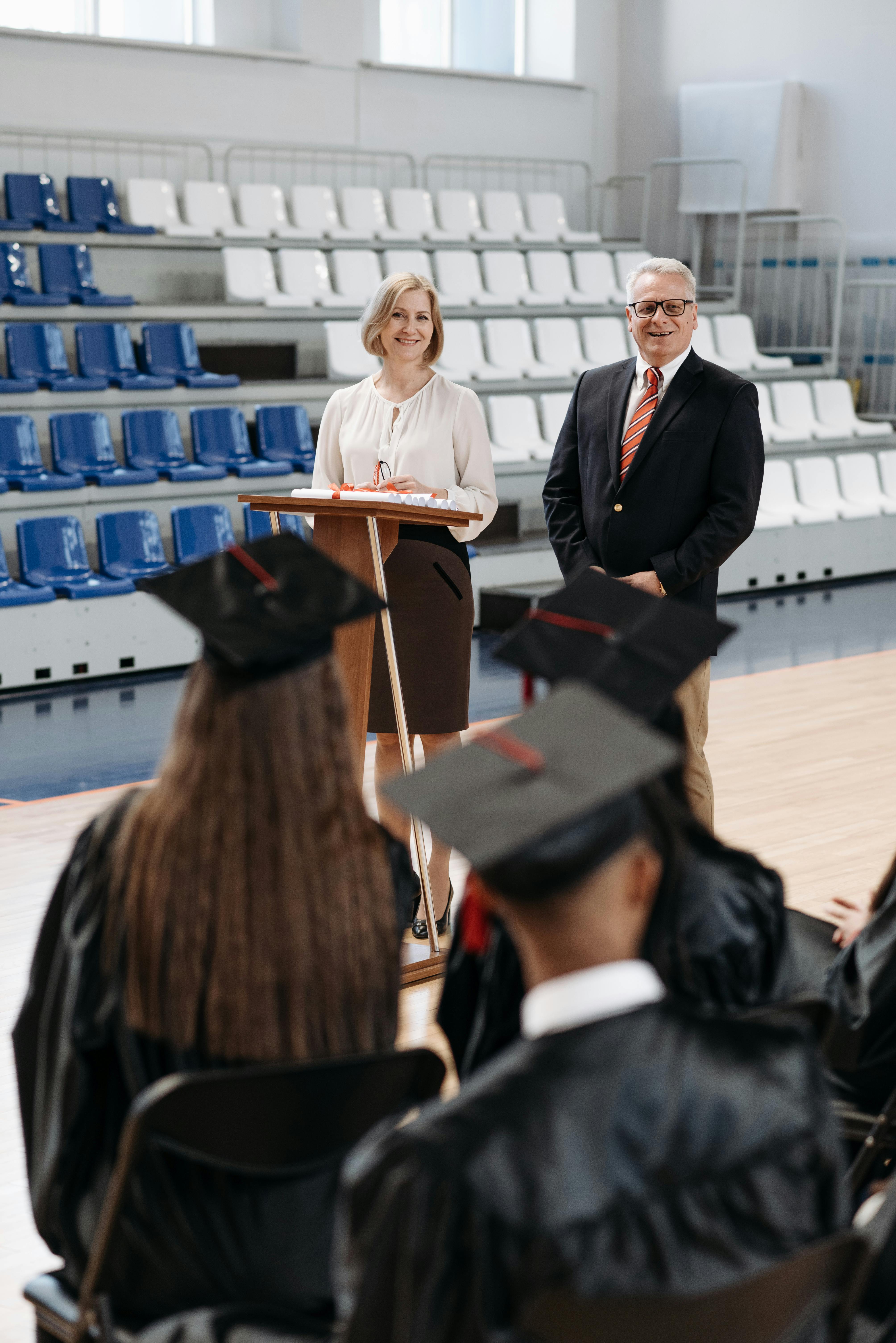 A formal graduation ceremony in a gymnasium with two speakers addressing graduates.